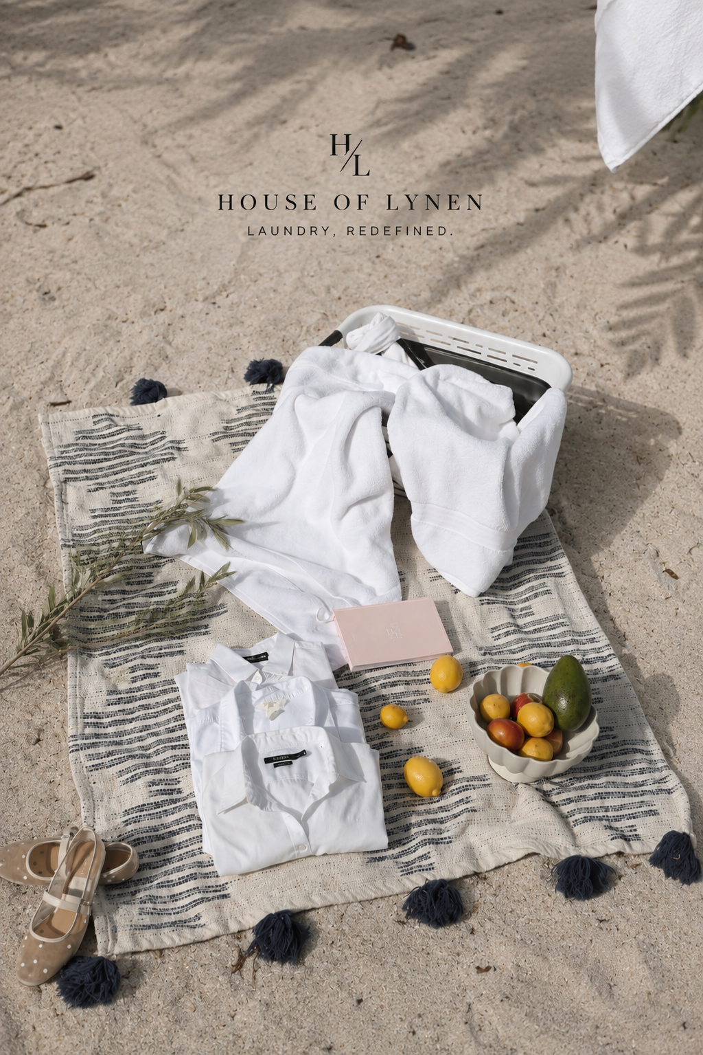 A sunlit beach flat lay of freshly folded white shirts and towels in a linen basket, styled with citrus and a bowl of fruit.